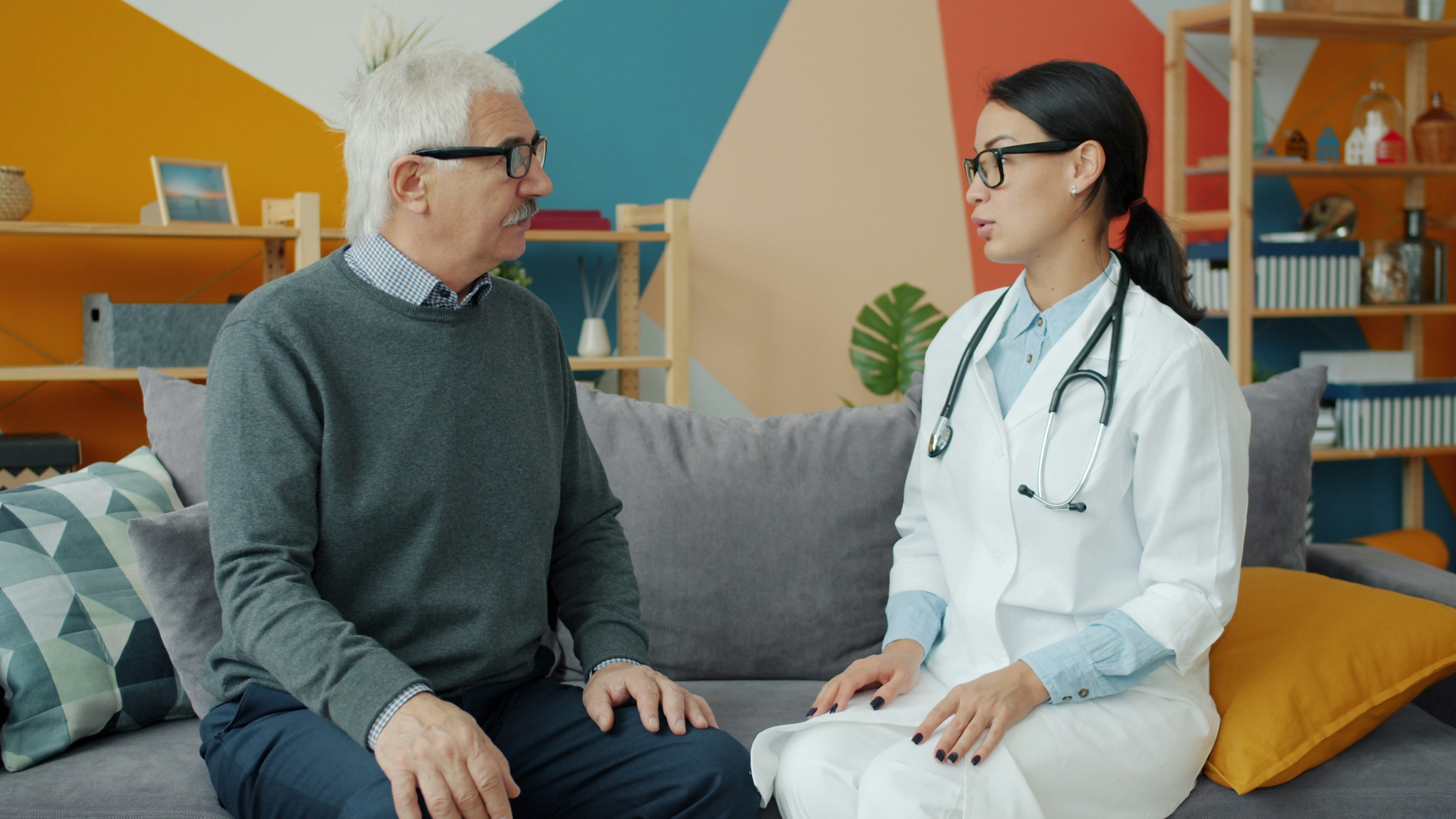 A physician and an older patient sit side by side on a sofa, talking in a bright, informal room